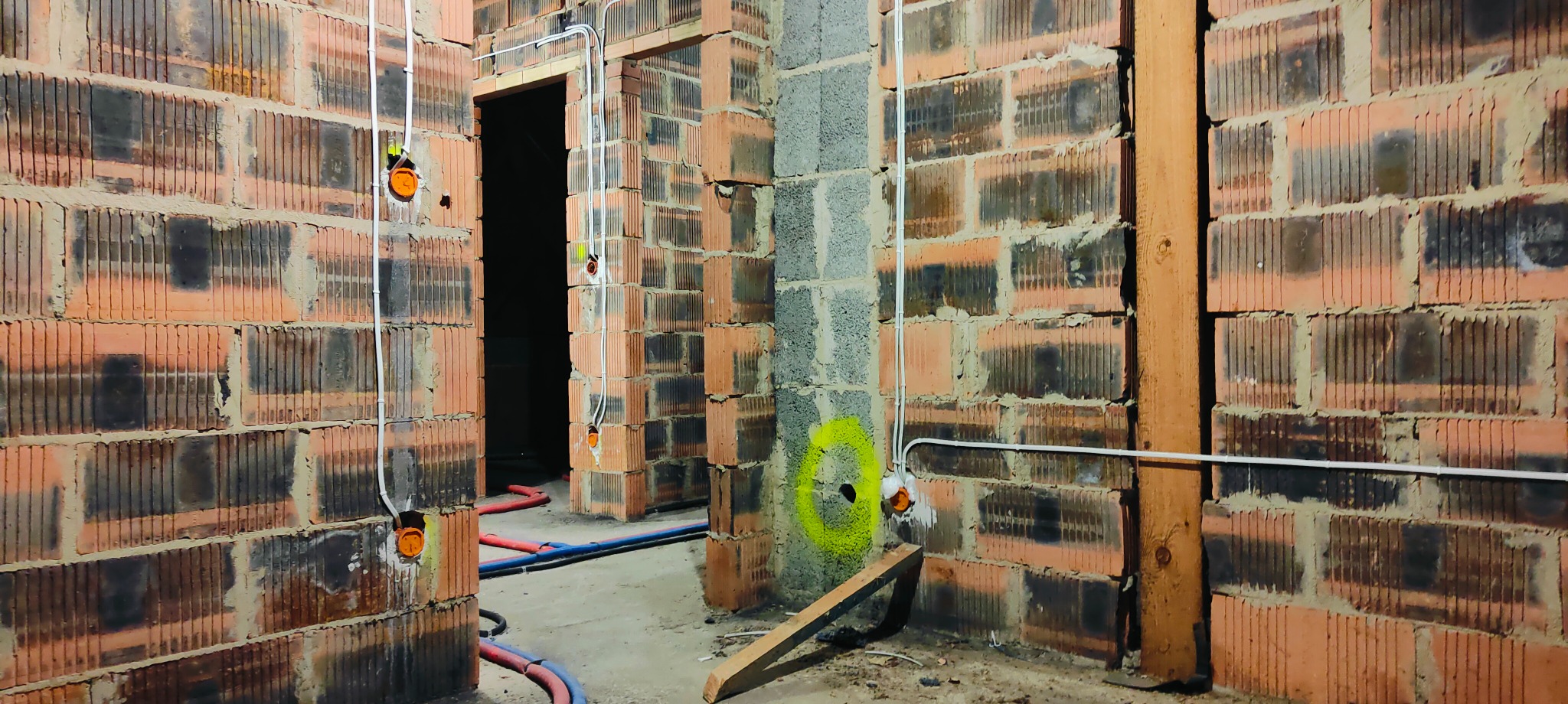Surowy mur z cegły dziurawki z widocznymi przewodami elektrycznymi w białych rurkach, pomarańczowe puszki instalacyjne, niebieskie i czerwone rury wodne na podłodze, fragmenty ściany z betonu...