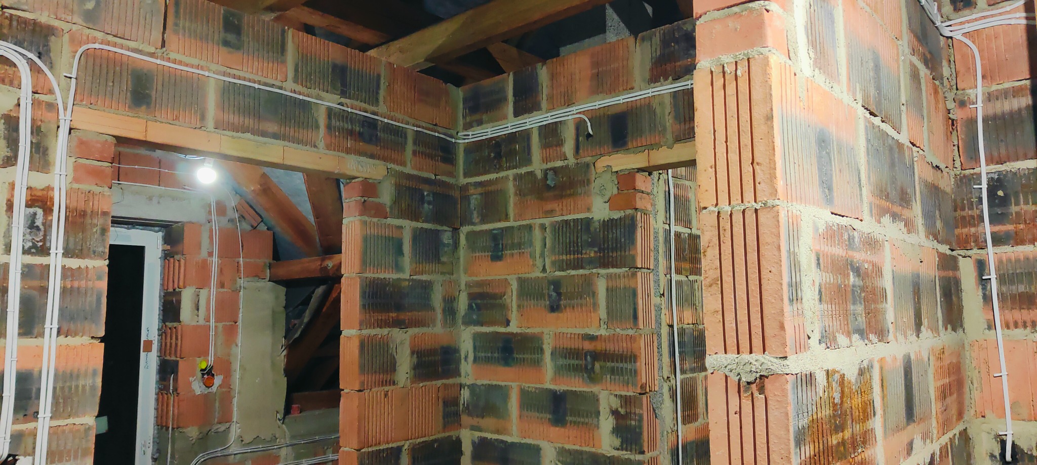 Wnętrze w trakcie prac budowlanych z widocznymi przewodami elektrycznymi w białych rurkach PCV, zamontowanymi na ścianach z czerwonej cegły dziurawki, pod sufitem widoczne drewniane belki stropowe,...