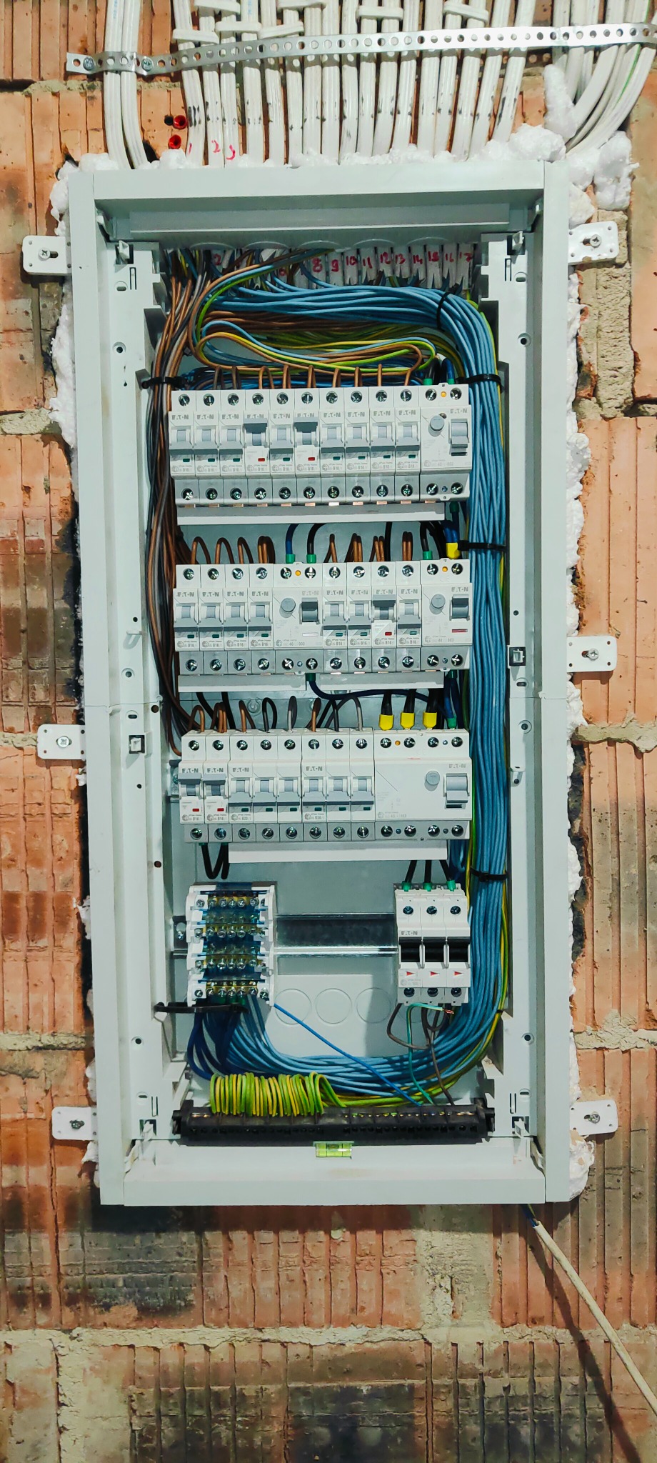 Rozdzielnica elektryczna z zabezpieczeniami, zamontowana na ścianie z czerwonej cegły, z widocznymi przewodami i elementami instalacji.