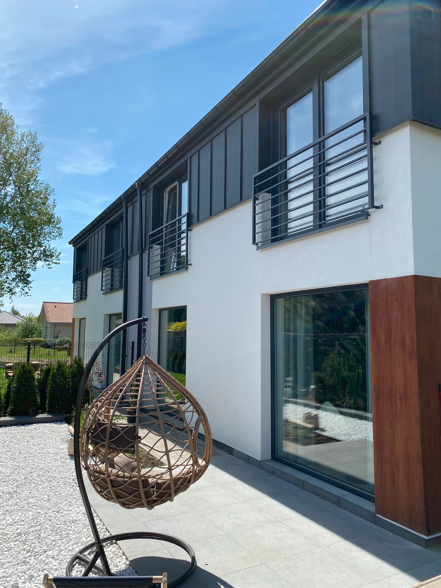 Nowoczesny dom z balkonami zabezpieczonymi czarnymi, metalowymi balustradami, widok z zewnątrz w słoneczny dzień, w tle białe kamienie i wiszące krzesło.