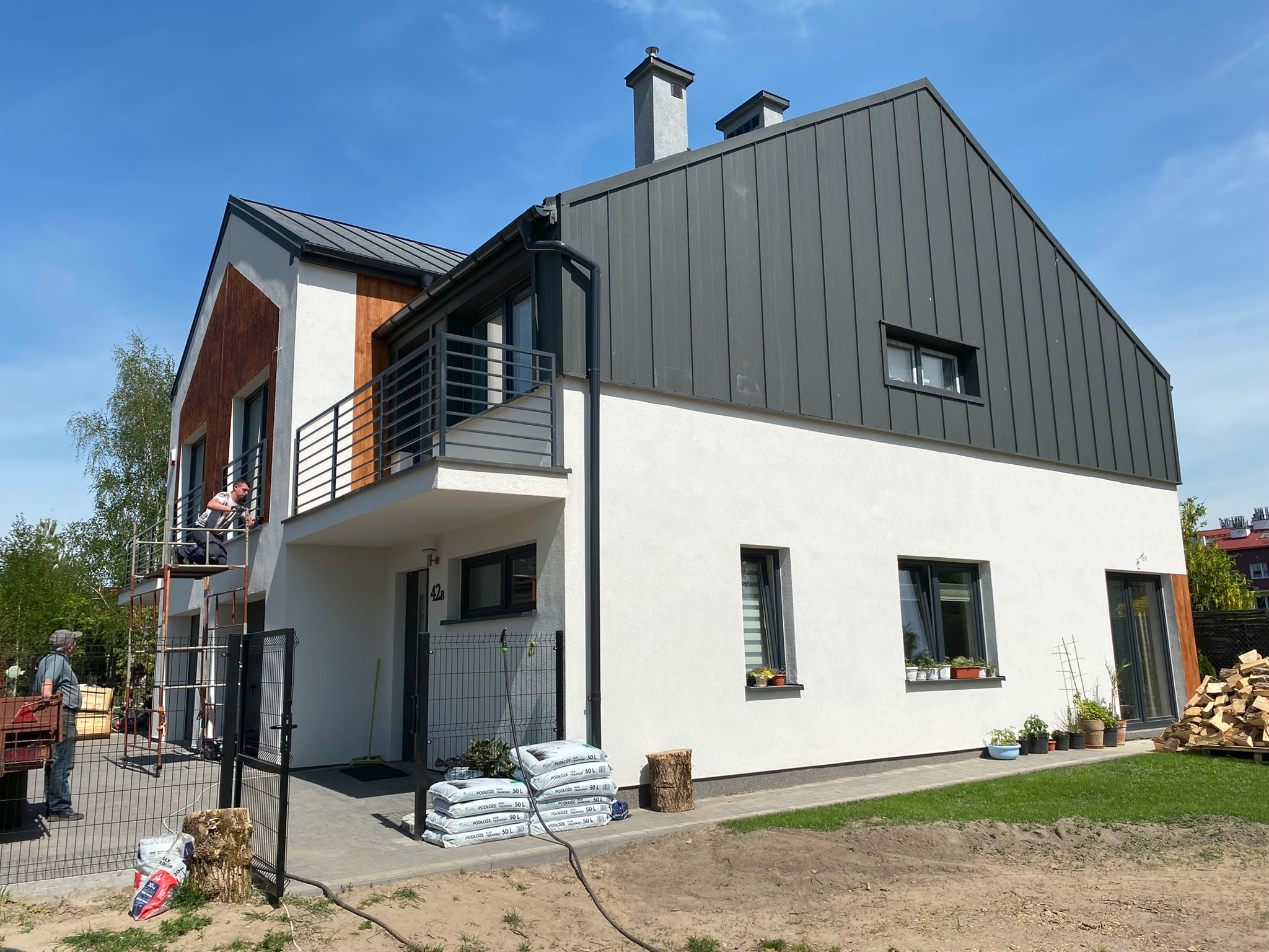 Montaż metalowej bramy wjazdowej na posesji, widoczny spawacz na rusztowaniu, brama częściowo zamontowana, worki z podłożem pod rośliny obok