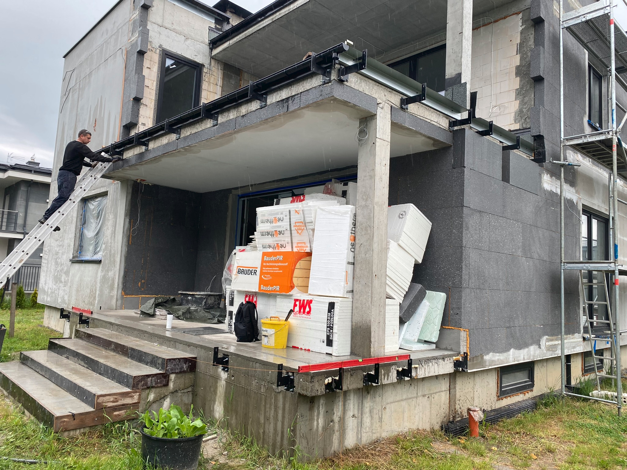 Prace wykończeniowe elewacji domu jednorodzinnego: robotnik na drabinie montuje orynnowanie, widoczne ocieplenie ścian styropianem i materiały izolacyjne na tarasie.