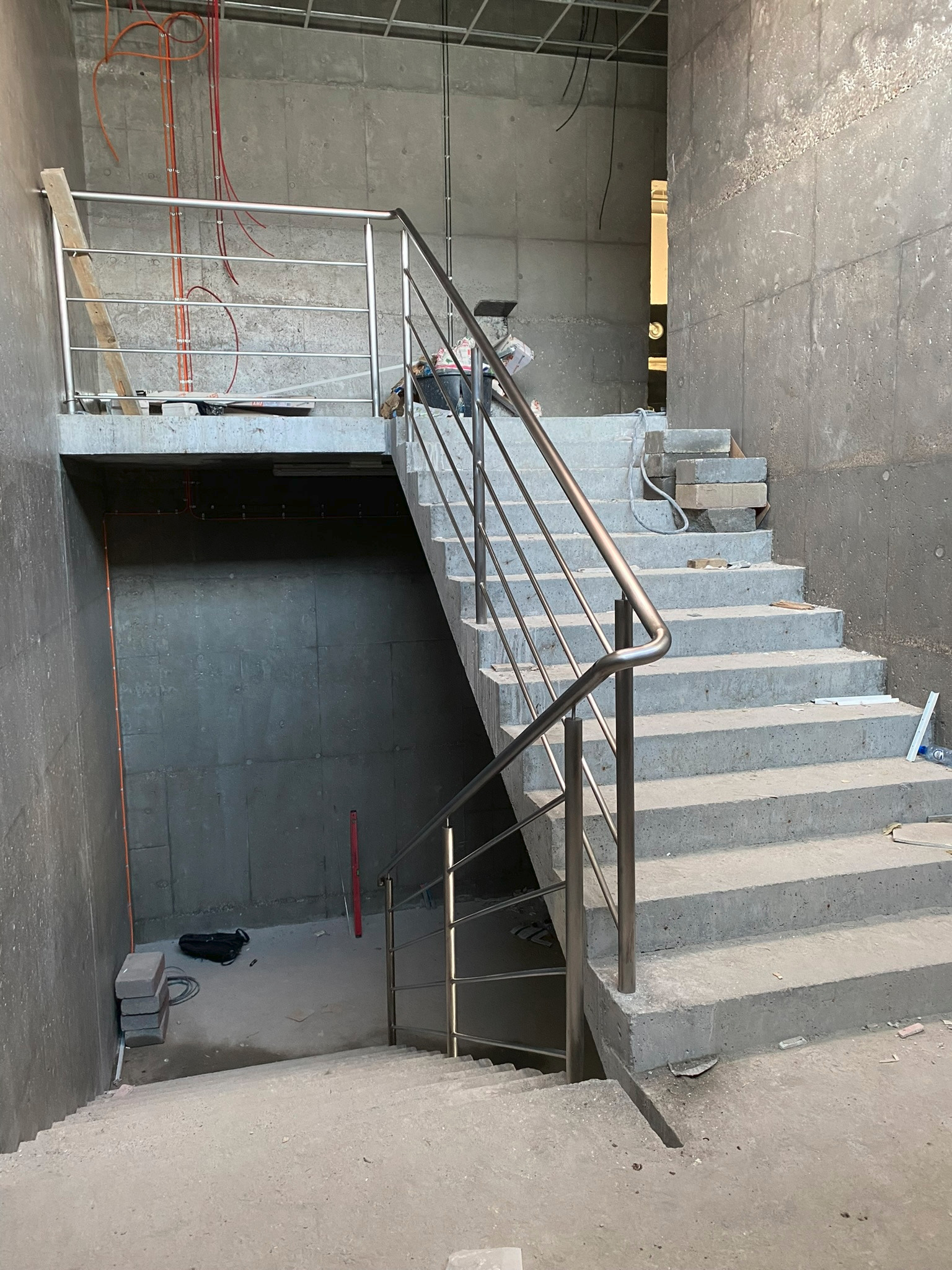 Surowe betonowe schody z zamontowaną, nowoczesną balustradą ze stali nierdzewnej w trakcie prac wykończeniowych, widoczne kable i materiały budowlane.