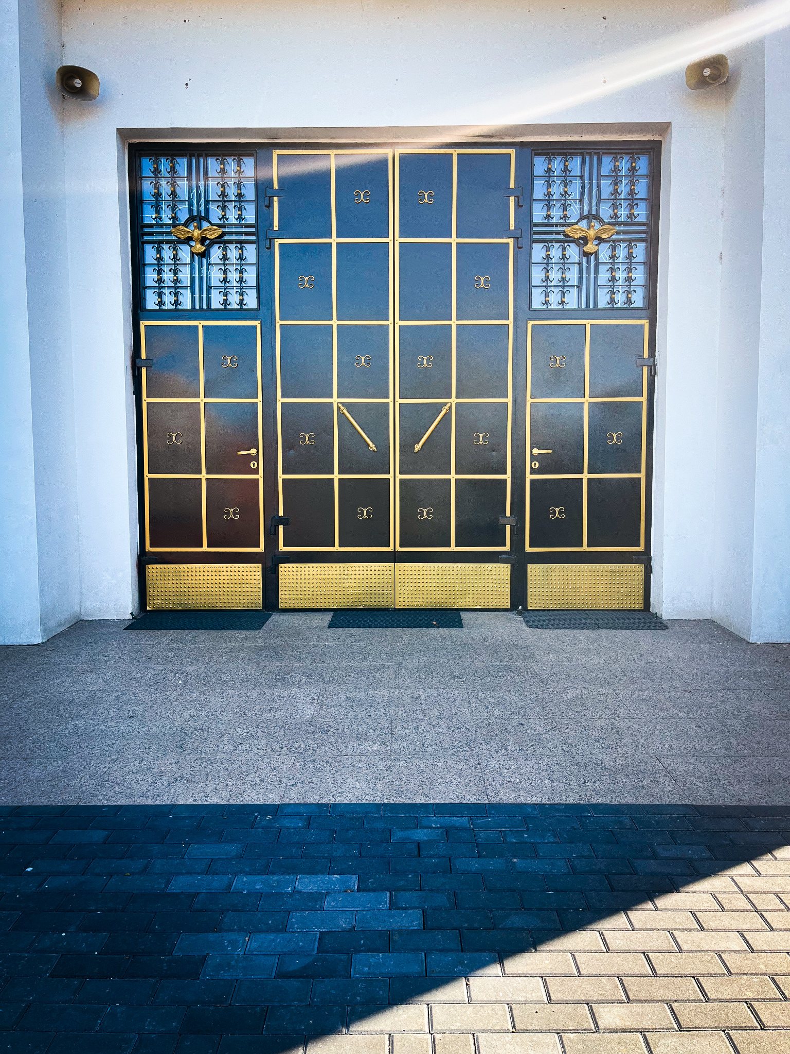 Czarna, metalowa brama wjazdowa z dekoracyjnymi, złotymi elementami kratownicy i godłem orła nad wejściem, umieszczona w białej fasadzie budynku.