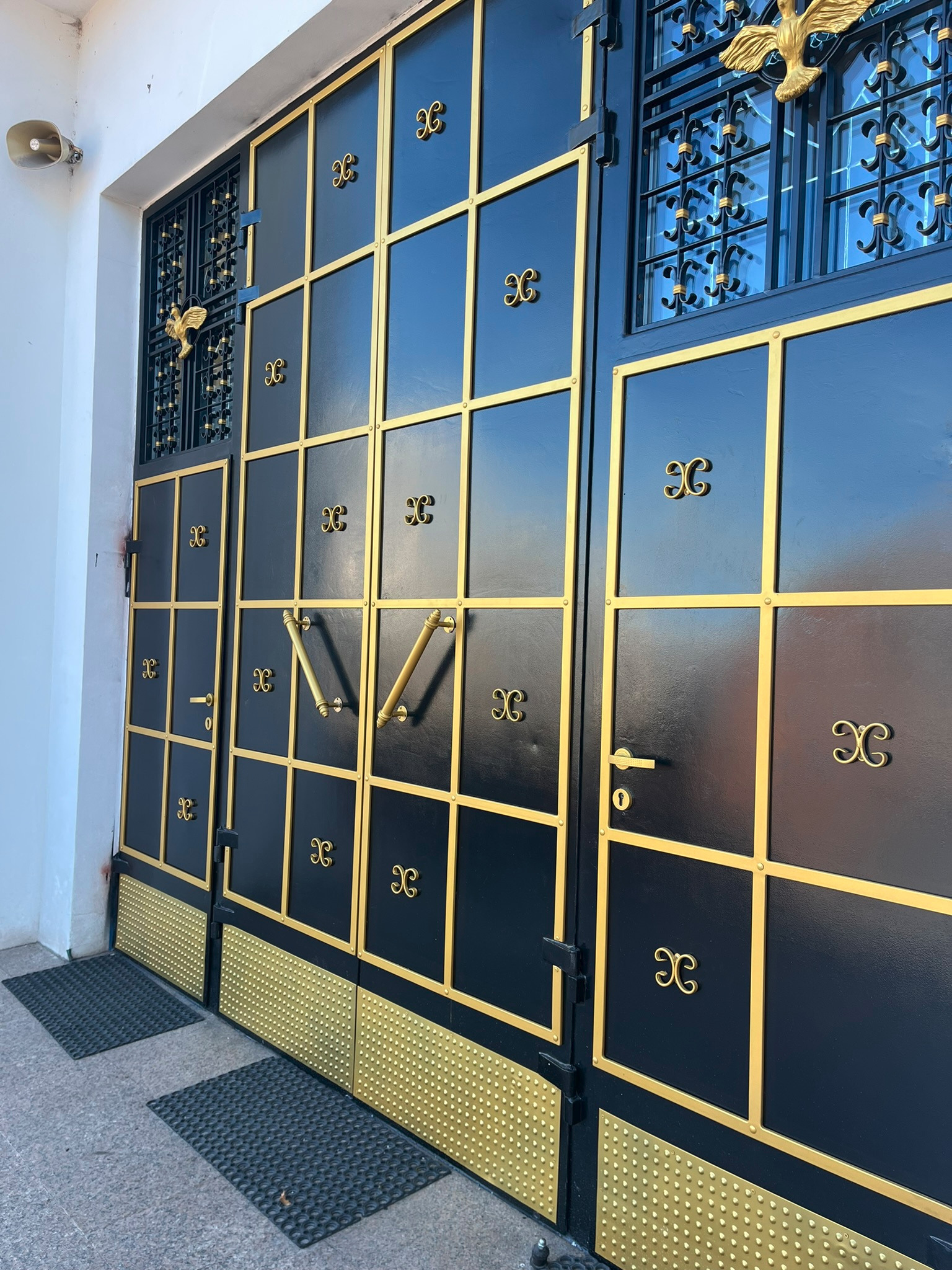 Czarna brama wjazdowa z geometrycznym złotym zdobieniem, widoczne detale jak uchwyty, zamek i dekoracyjne elementy, na tle białej ściany budynku, z wycieraczkami przed bramą.