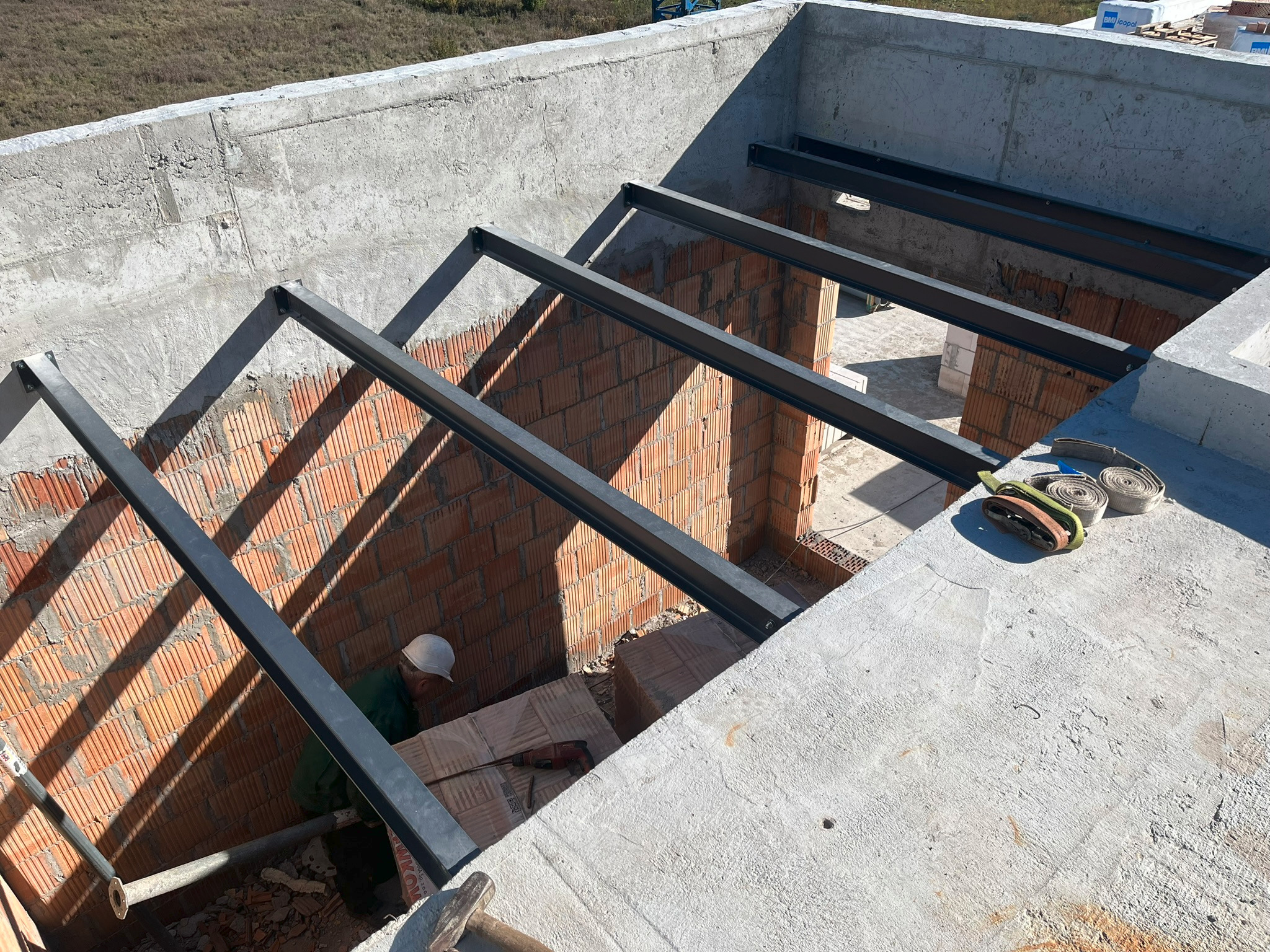 Konstrukcja metalowa dachu w trakcie budowy, widok z góry, ceglane ściany, pracownik w kasku przy pracy, betonowe elementy konstrukcyjne.