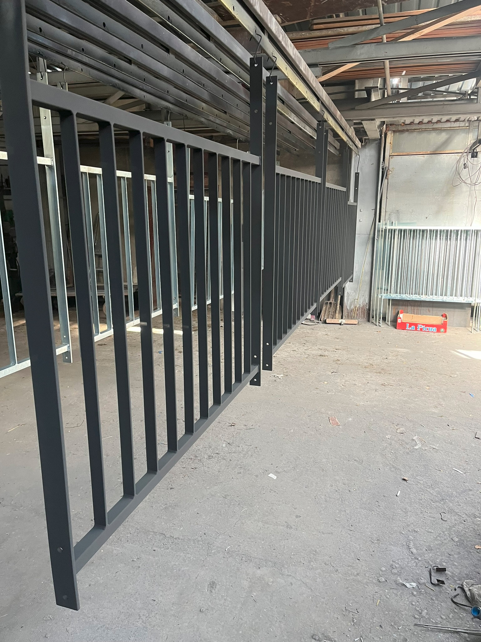 Szara metalowa balustrada w trakcie malowania proszkowego, zawieszona na wieszakach w hali produkcyjnej, widoczna podłoga hali i karton po owocach.