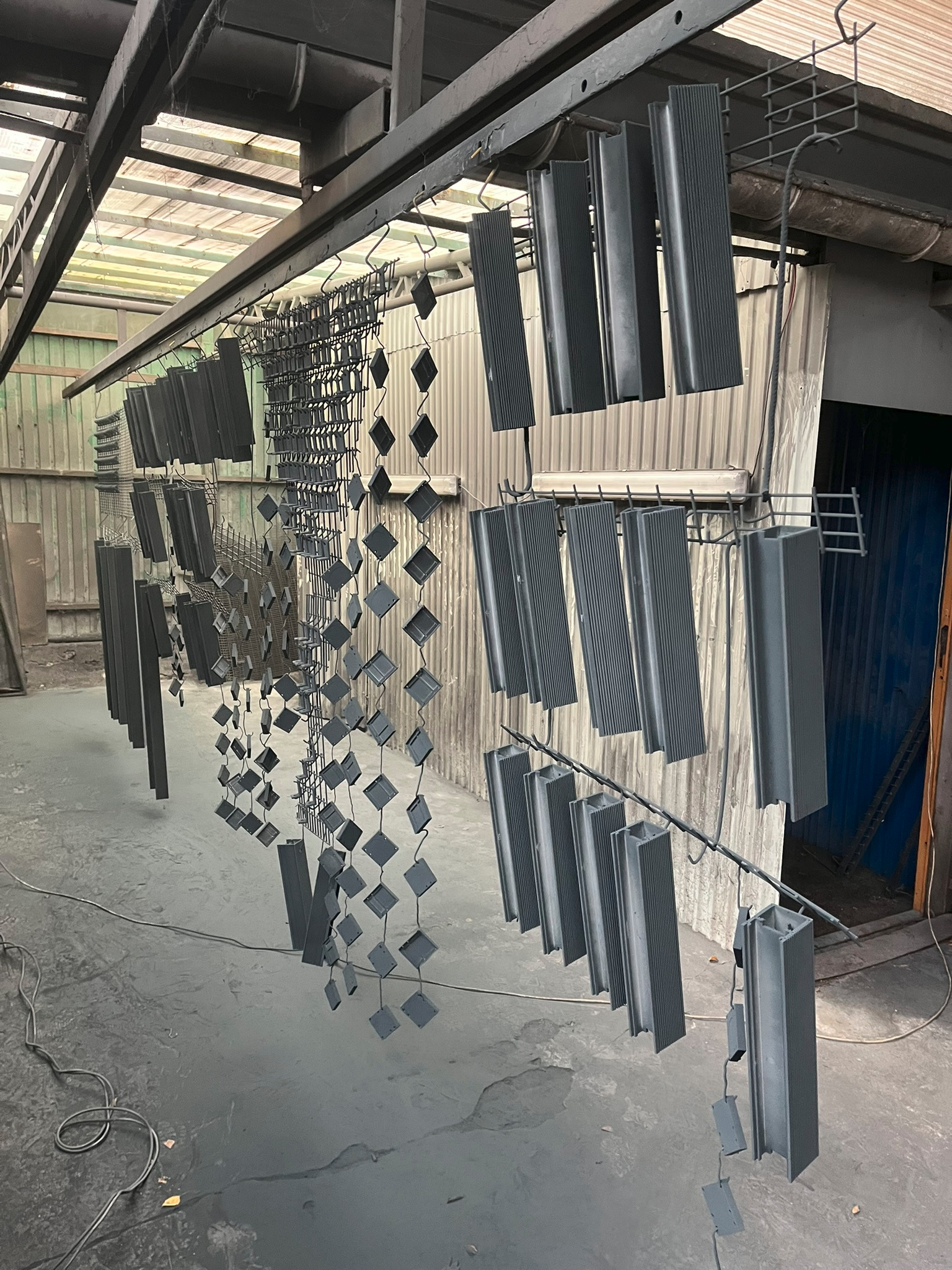 Elementy metalowe o różnych kształtach, prawdopodobnie do ogrodzeń lub bram, zawieszone na wieszakach po malowaniu proszkowym w szarej hali produkcyjnej.