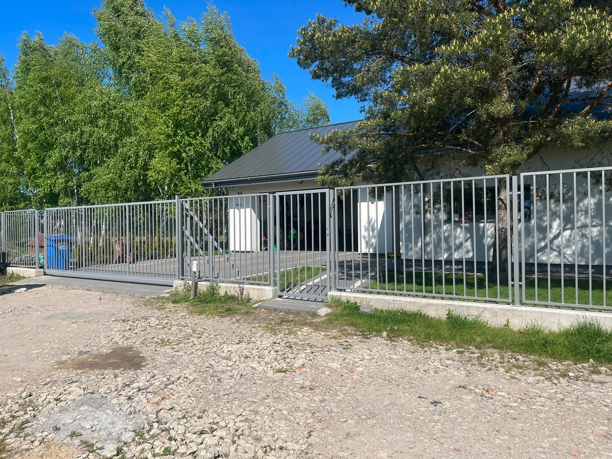Szara przesuwna brama wjazdowa z metalowymi sztachetami, widok z zewnątrz na żwirowy podjazd, z budynkiem w tle i zielenią drzew.