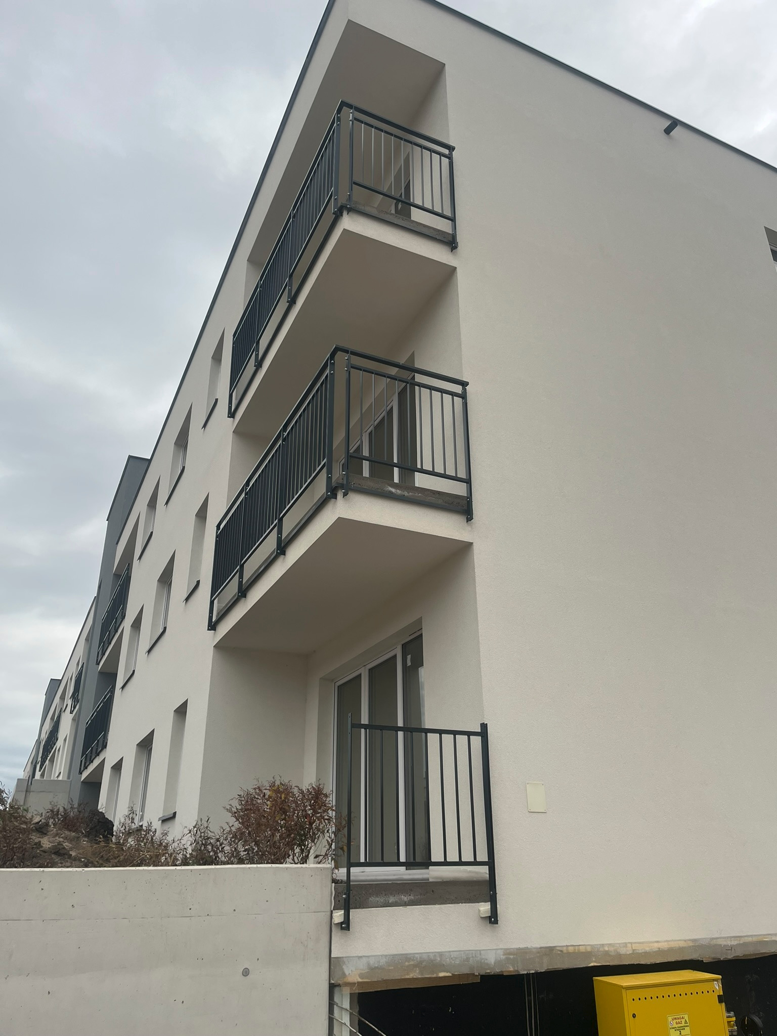 Nowoczesny budynek wielorodzinny z balkonami zabezpieczonymi czarnymi, metalowymi barierkami, widok z dołu na jasną elewację i pochmurne niebo.
