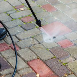 Grzegorz Gańczak - Czyszczenie kostki brukowej myjką ciśnieniową. Dysza spryskuje czerwoną kostkę, w tle widoczne fragmenty niebieskiego kombinezonu i butów pracownika.