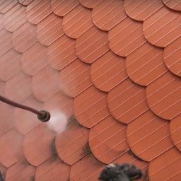 Grzegorz Gańczak - Czerwona dachówka łuskowa czyszczona myjką ciśnieniową. Woda spływa do czarnej rynny. Fragment białego samochodu w tle. Widok z góry.