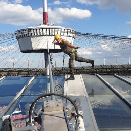 Grzegorz Gańczak - Pracownik w kasku i uprzęży, balansuje na dachu stadionu, prawdopodobnie podczas prac konserwacyjnych lub inspekcyjnych. Widoczne szklane panele dachu i konstrukcja nośna.