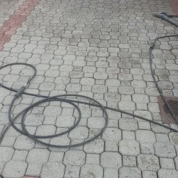 Częściowo umyta kostka brukowa z widocznym wężem ciśnieniowym i śladami wody, ukazująca efekt czyszczenia. Fragment ogrodzenia i budynku w tle.