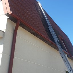 Czerwony dach skośny z rynną i drabiną opartą o niego, na tle błękitnego nieba. Widoczna jasna elewacja budynku z małym oknem.