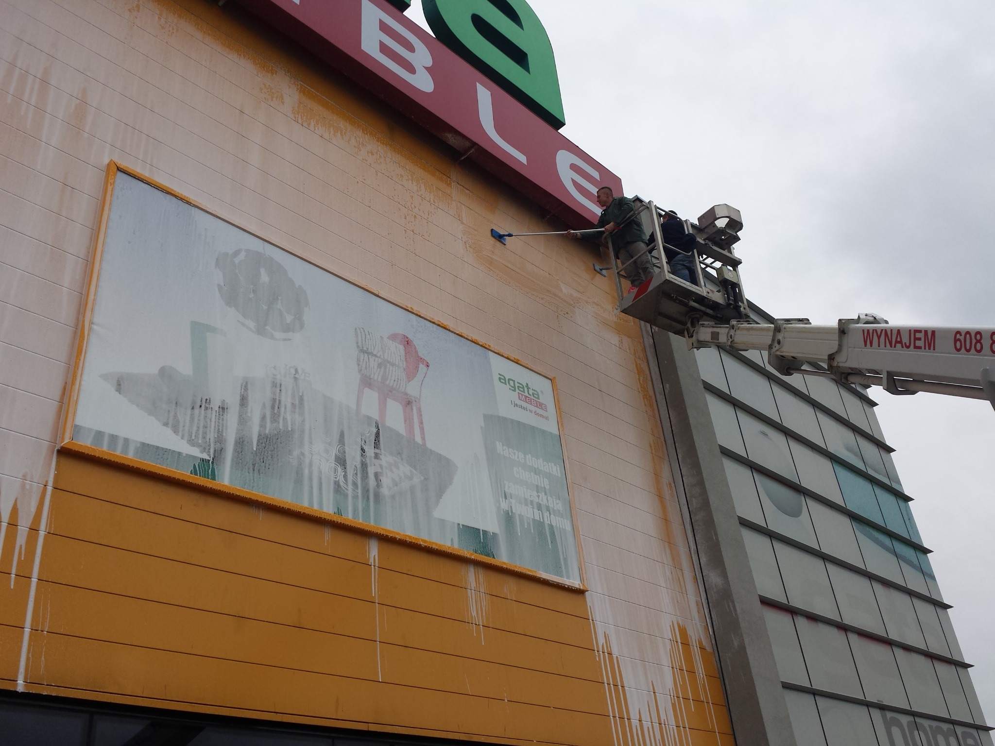 Pracownik na podnośniku czyści elewację budynku handlowego z reklamą Agata Meble. Widoczne zacieki po myciu i narzędzie do czyszczenia na długim kiju.