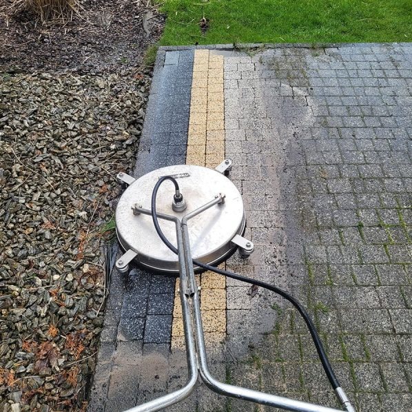 Mycie kostki brukowej urządzeniem ciśnieniowym. Widoczna różnica między czystą a zabrudzoną powierzchnią. Częściowo widoczne otoczenie z kamyków i trawy.