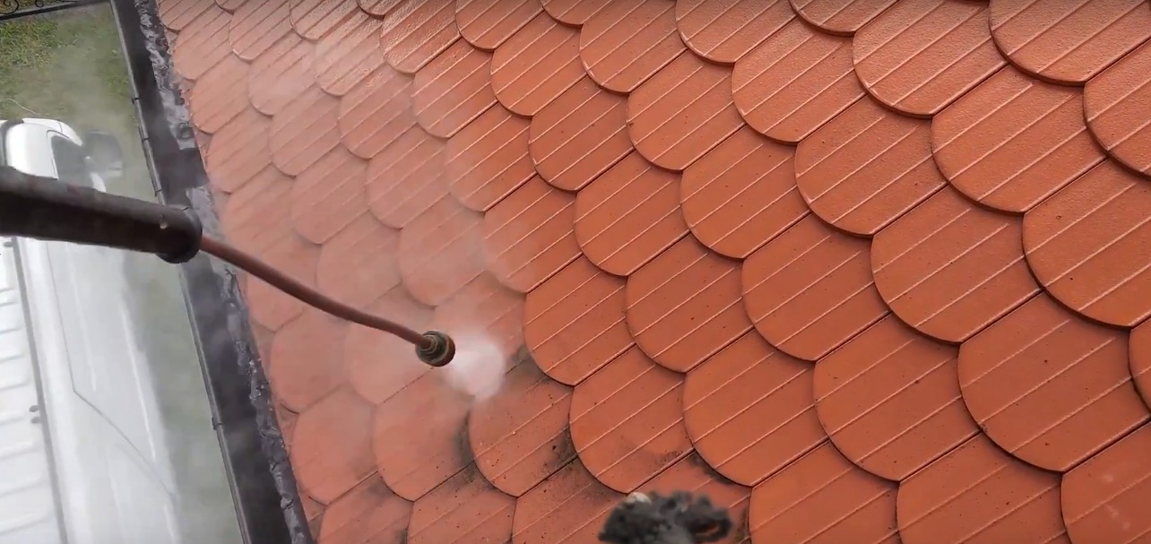 Czerwona dachówka łuskowa czyszczona myjką ciśnieniową. Woda spływa do czarnej rynny. Fragment białego samochodu w tle. Widok z góry.
