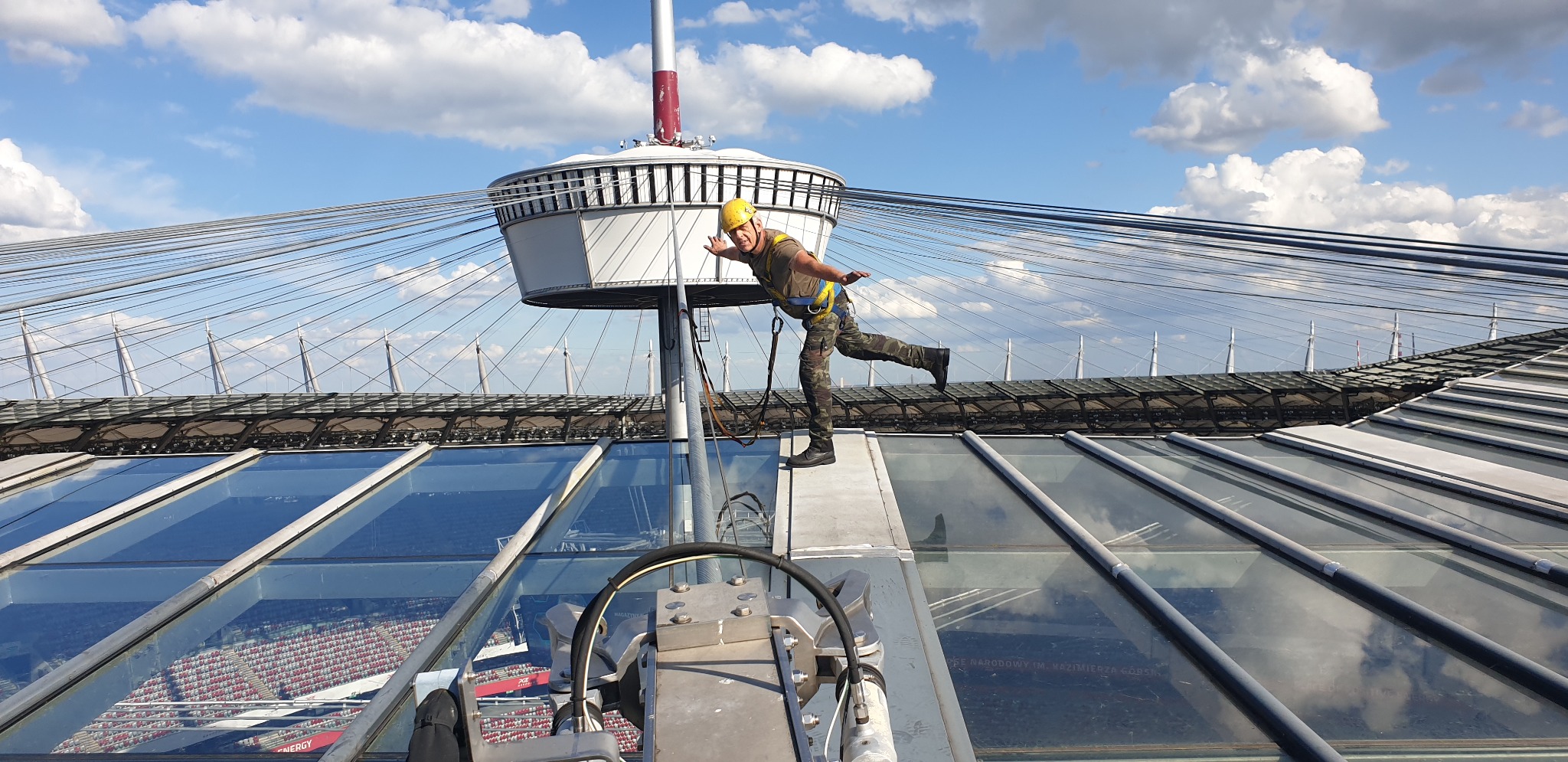 Pracownik w kasku i uprzęży, balansuje na dachu stadionu, prawdopodobnie podczas prac konserwacyjnych lub inspekcyjnych. Widoczne szklane panele dachu i konstrukcja nośna.