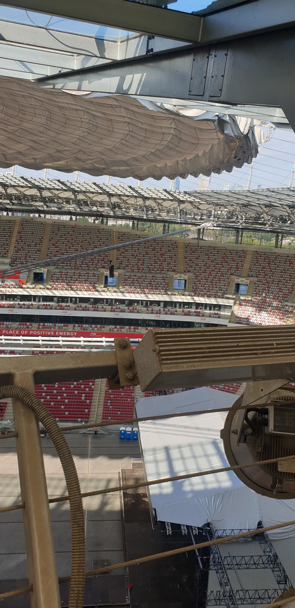 Widok z góry na stadion z elementami konstrukcyjnymi i sprzętem. W tle widoczne trybuny i zadaszenie stadionu. Ujęcie z dużej wysokości.