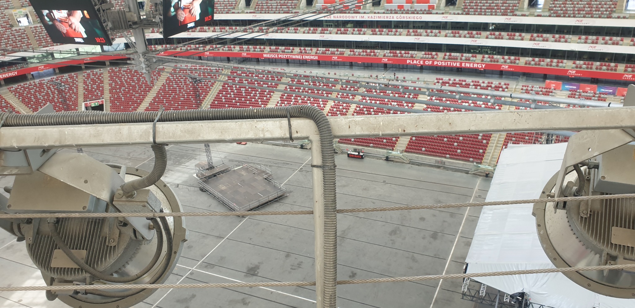 Widok z góry na stadion w Piotrków Kujawski z widocznymi reflektorami i sceną. Perspektywa z wysokości, ukazująca trybuny i plac. Elementy konstrukcyjne na pierwszym planie.