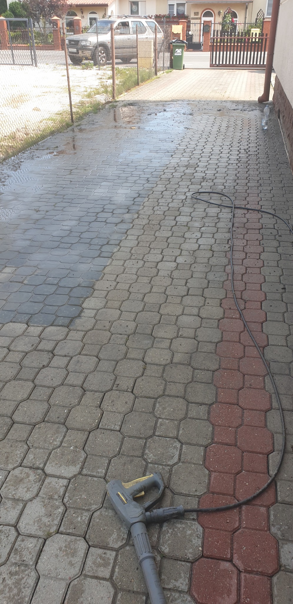 Częściowo umyta kostka brukowa na podjeździe, widoczna różnica w kolorze. W tle dom, ogrodzenie i samochód. Na pierwszym planie lanca myjki ciśnieniowej.