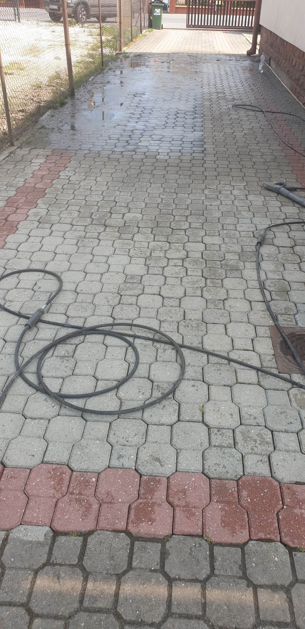 Częściowo umyta kostka brukowa z widocznym wężem ciśnieniowym i śladami wody, ukazująca efekt czyszczenia. Fragment ogrodzenia i budynku w tle.