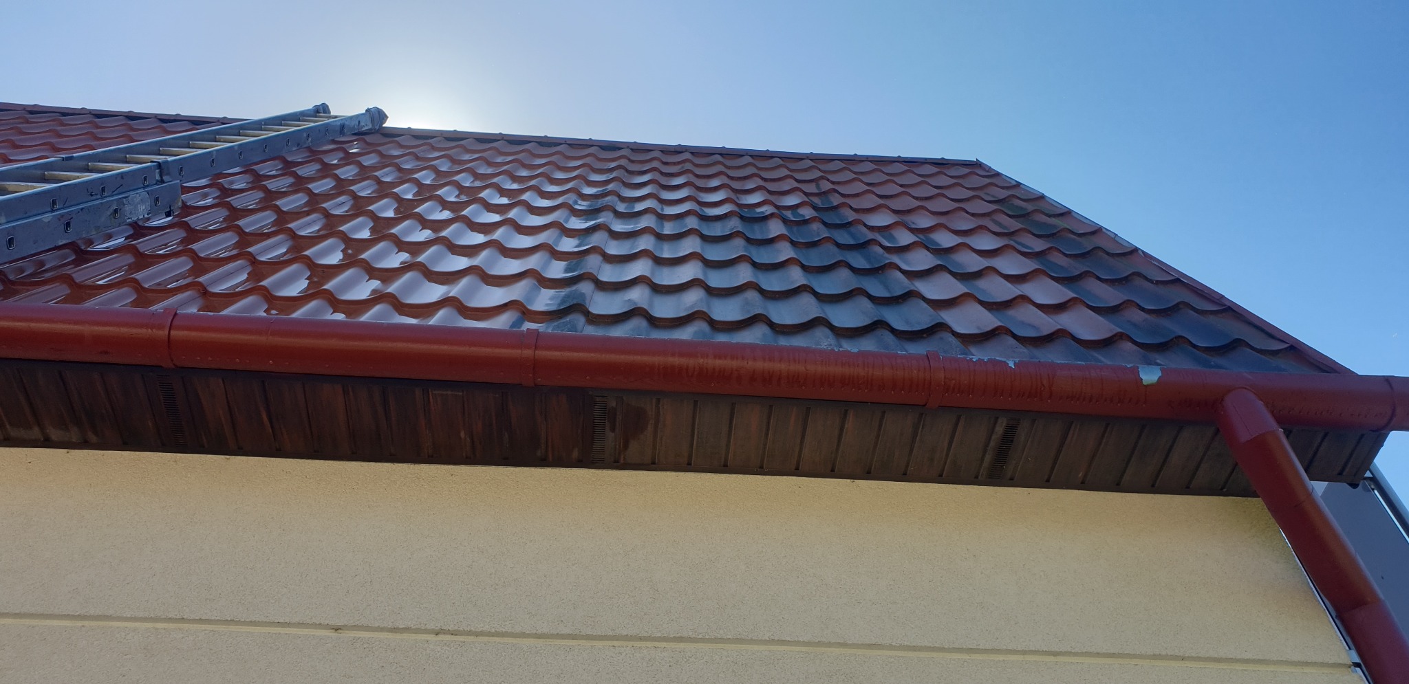 Częściowo umyty, błyszczący dach z metalowej dachówki w kolorze ceglastym, z widoczną drabiną i rynną, na tle błękitnego nieba.