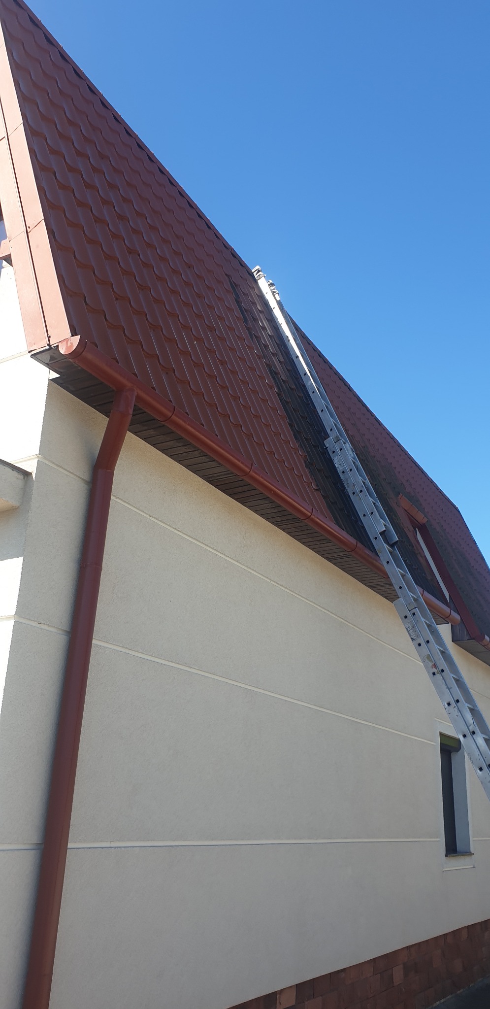 Czerwony dach skośny z rynną i drabiną opartą o niego, na tle błękitnego nieba. Widoczna jasna elewacja budynku z małym oknem.