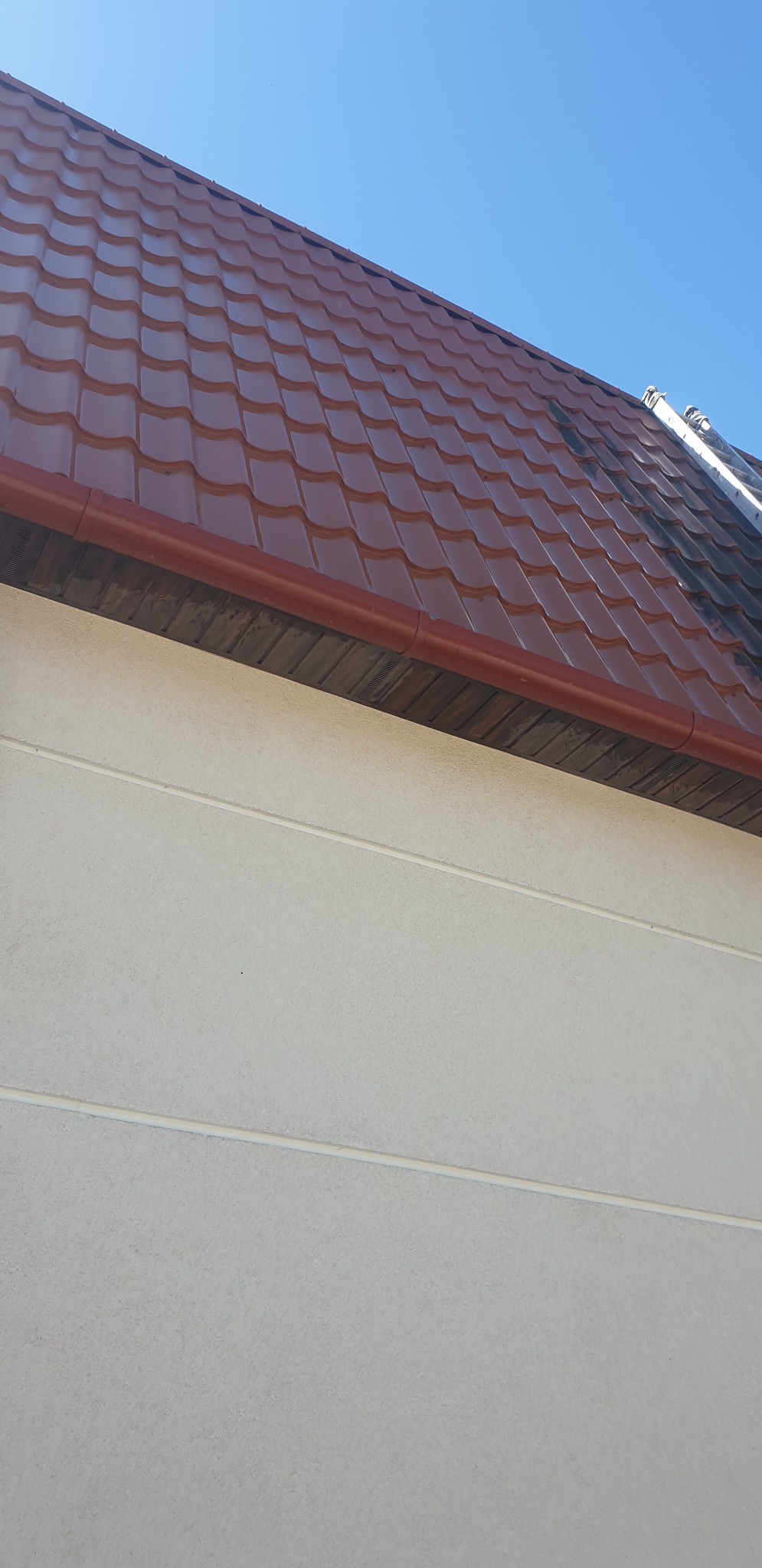 Czerwono-brązowy dach z blachodachówki, jasna elewacja budynku i fragment błękitnego nieba. Widoczny fragment rynny i drabiny na dachu.