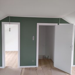 Krzysztof Borowiec - Wnętrze w trakcie prac: ściana pomalowana na oliwkowy kolor, białe futryny drzwiowe, surowe podłoże pod panele zabezpieczone kartonem, widoczne gniazdka i wisząca żarówka.