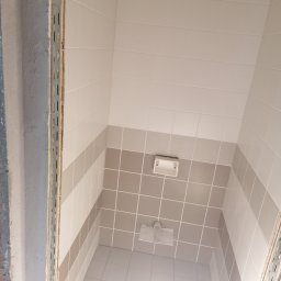 Krzysztof Borowiec - Wnętrze łazienki w trakcie remontu, widoczne płytki ścienne w odcieniach bieli i szarości, fragment sufitu z instalacją oraz metalowa listwa przy ościeżnicy.