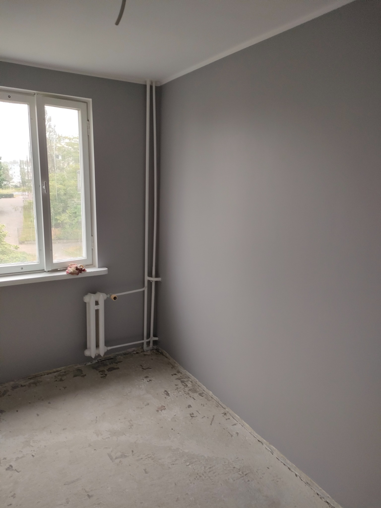 Pomieszczenie pomalowane na szaro z białym oknem i grzejnikiem, podłoga w trakcie przygotowania do dalszych prac wykończeniowych.