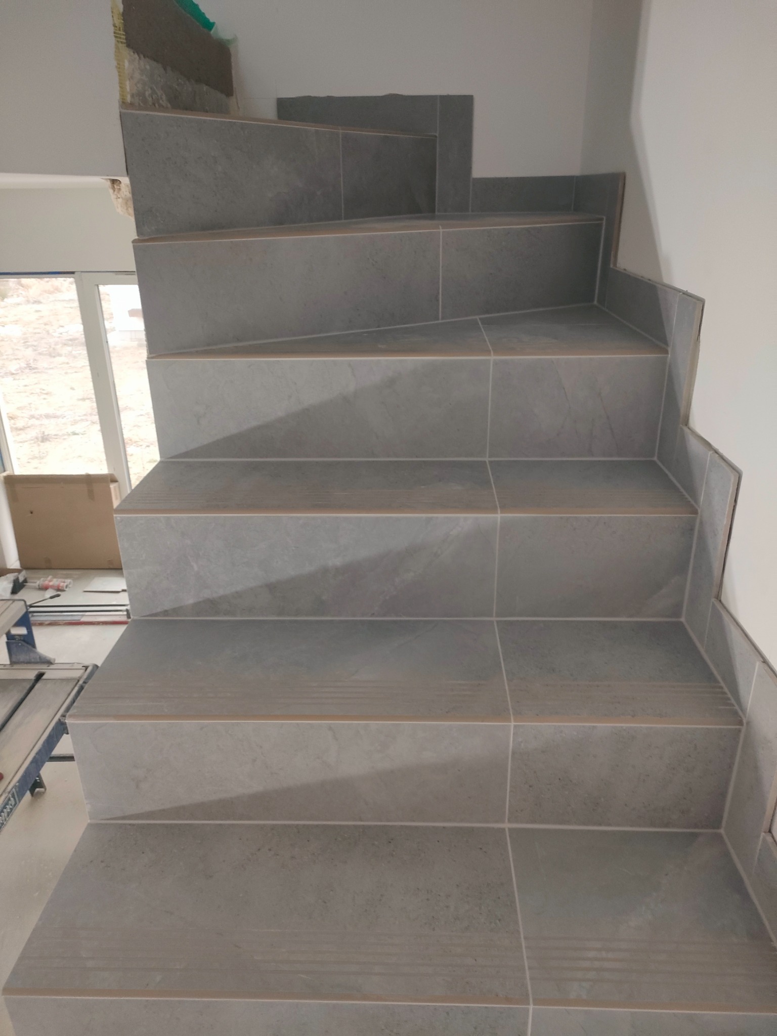 Wykończone schody wewnętrzne wyłożone szarymi płytkami, widoczne narzędzia glazurnicze i karton w tle, fragment surowego wykończenia ściany przy schodach.