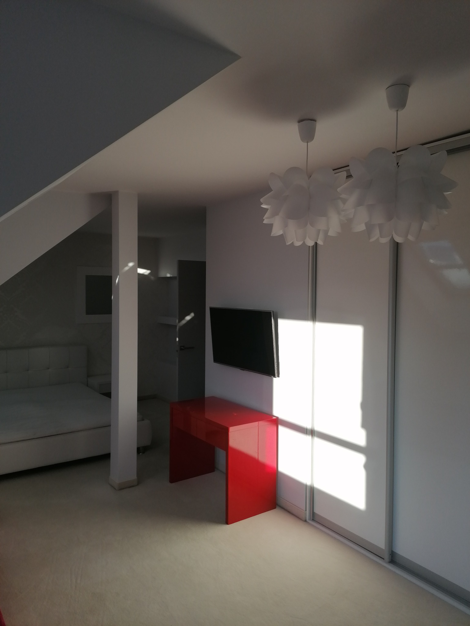 Jasne wnętrze pokoju z białymi ścianami, szafą przesuwną, czerwonym biurkiem pod telewizorem i dwoma białymi lampami w kształcie kwiatów