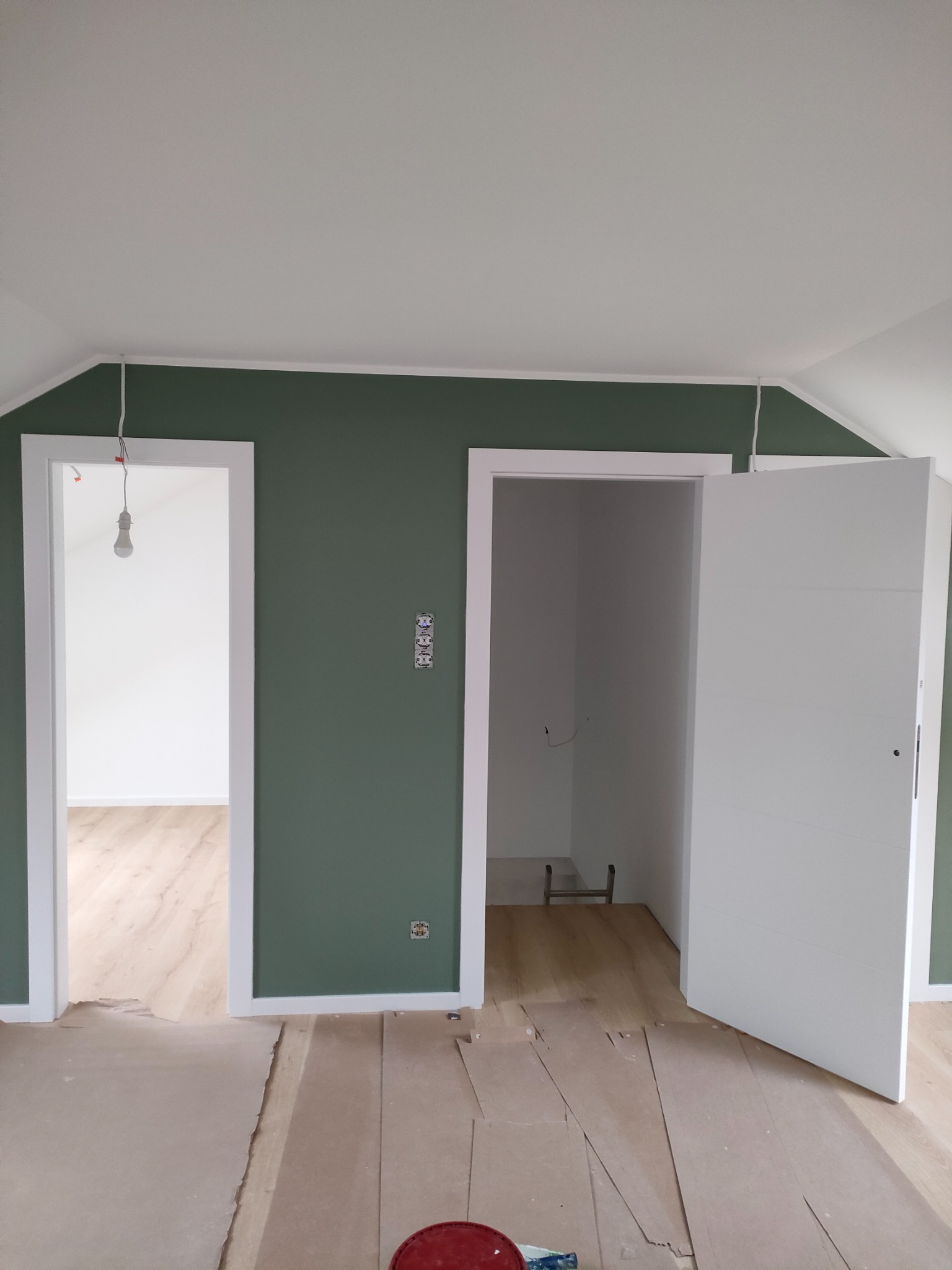 Wnętrze w trakcie prac: ściana pomalowana na oliwkowy kolor, białe futryny drzwiowe, surowe podłoże pod panele zabezpieczone kartonem, widoczne gniazdka i wisząca żarówka.