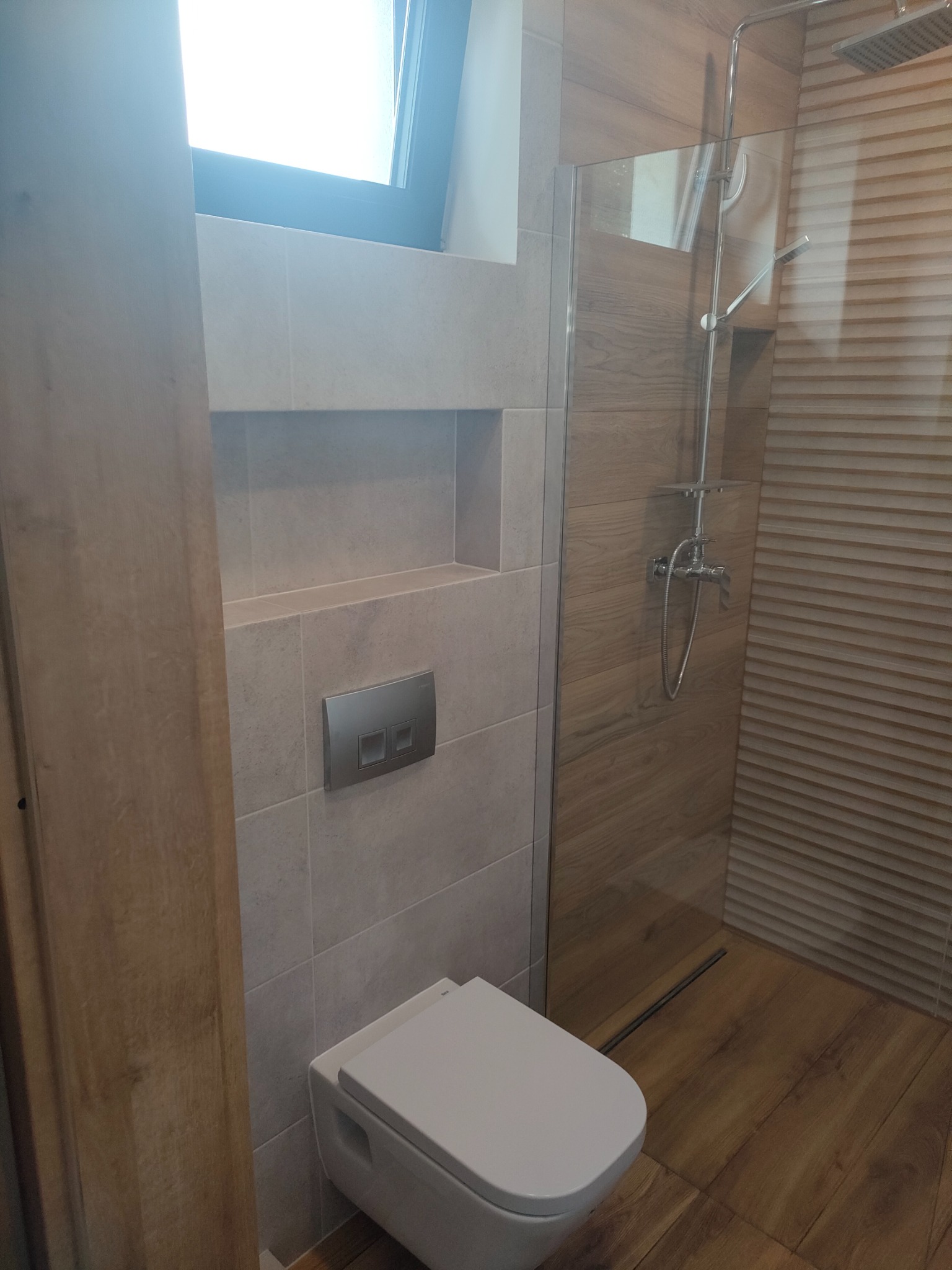 Nowoczesna łazienka z drewnopodobnymi płytkami, podwieszana toaleta z chromowanym przyciskiem spłukiwania, kabina prysznicowa ze szklaną ścianką i deszczownicą.