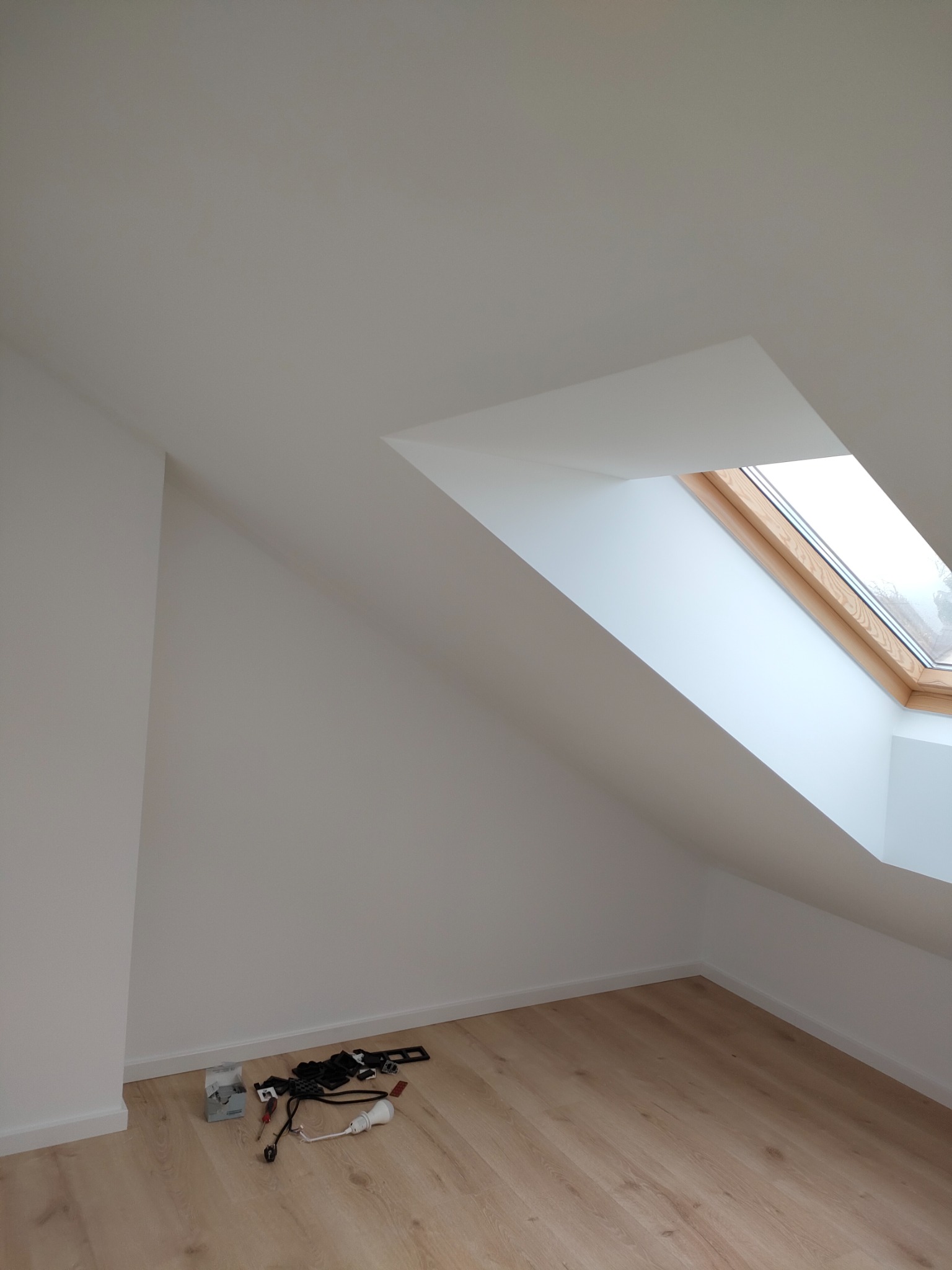 Poddasze po remoncie: świeżo pomalowane ściany, jasna podłoga panelowa, okno dachowe z drewnianą ramą, narzędzia i akcesoria do montażu oświetlenia leżące przy ścianie.