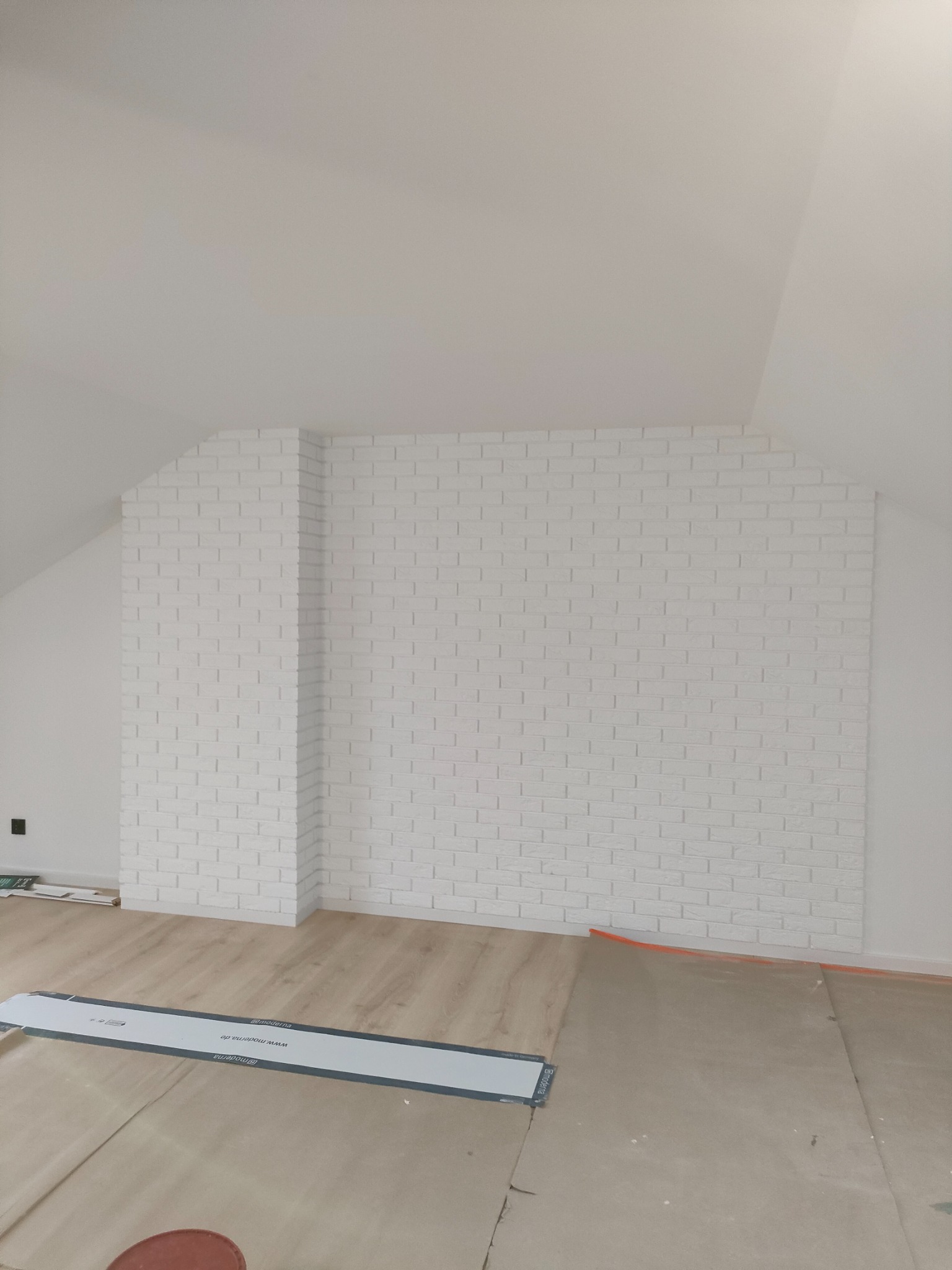Ściana w pokoju na poddaszu wykończona białą cegłą dekoracyjną, widoczne fragmenty podłogi w trakcie zabezpieczania przed dalszymi pracami.