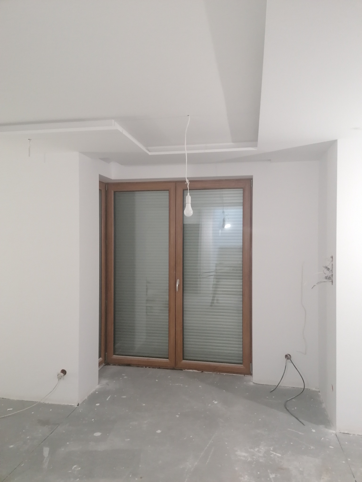Pokój w trakcie remontu: białe ściany po szpachlowaniu, sufit z elementami z płyty karton gips, niepodłączone kable elektryczne, drewniane okno z żaluzjami.