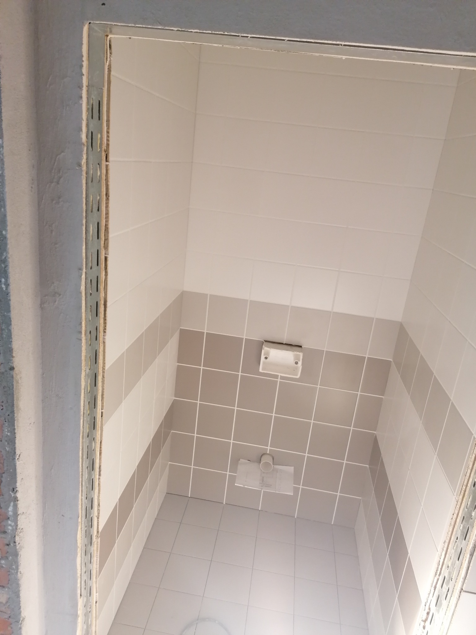 Wnętrze łazienki w trakcie remontu, widoczne płytki ścienne w odcieniach bieli i szarości, fragment sufitu z instalacją oraz metalowa listwa przy ościeżnicy.
