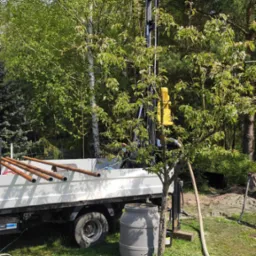 Wiertnica zamontowana na samochodzie ciężarowym na trawiastym terenie, widoczne rury wiertnicze, plastikowa beczka i drzewa w tle.