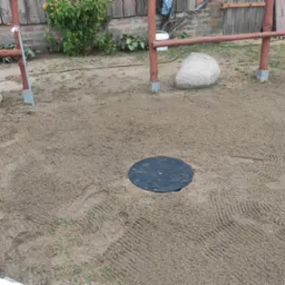 Teren po wykonaniu prac ziemnych z widoczną czarną pokrywą studzienki w centralnej części, otoczoną piaskiem z wyraźnymi śladami opon i butów, w tle drewniana konstrukcja i kamienie.