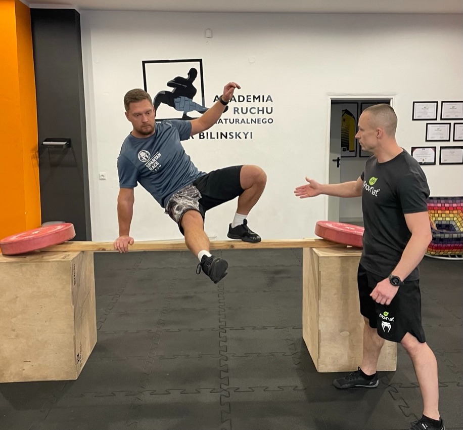 Mężczyzna w koszulce Spartan Race pokonuje przeszkodę na drewnianej belce, drugi mężczyzna w czarnej koszulce z logo stoi obok, wyciągając rękę w geście pomocy, w tle logo Akademii Ruchu Naturalnego.