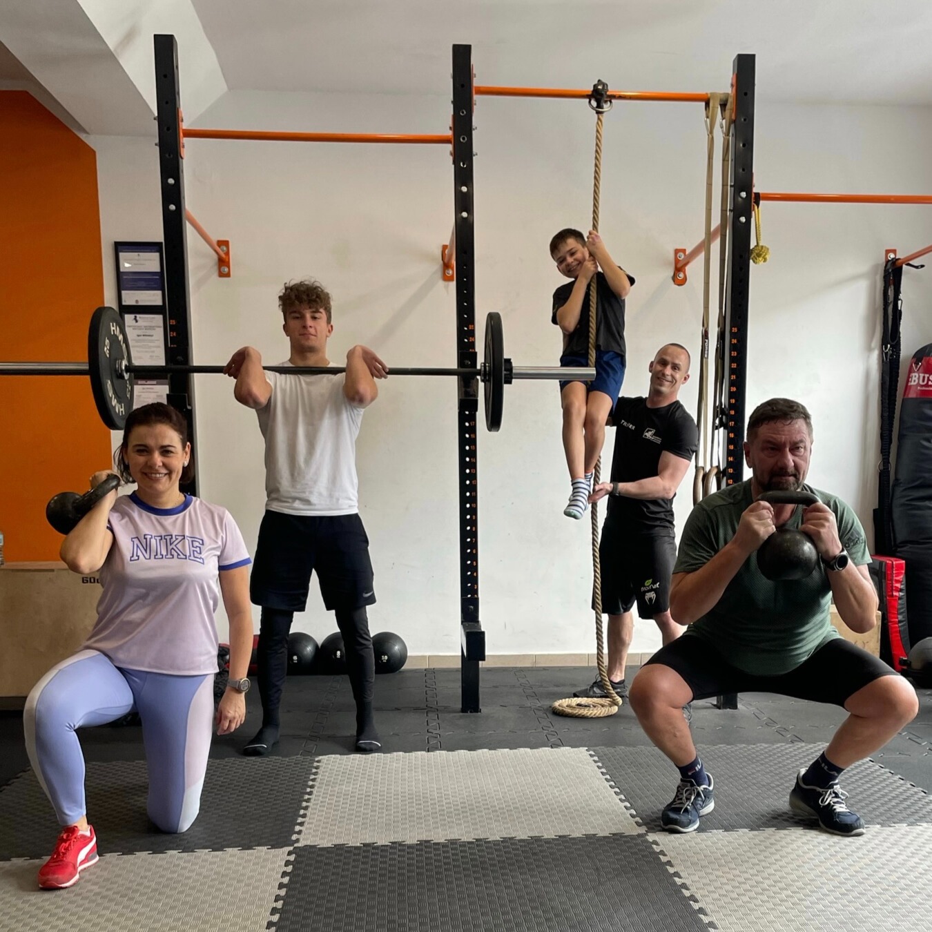 Grupa osób ćwiczących w siłowni: kobieta klęczy z kettlebellem, mężczyzna robi przysiad z kettlebellem, nastolatek podnosi sztangę, chłopiec wspina się po linie, a trener asekuruje.