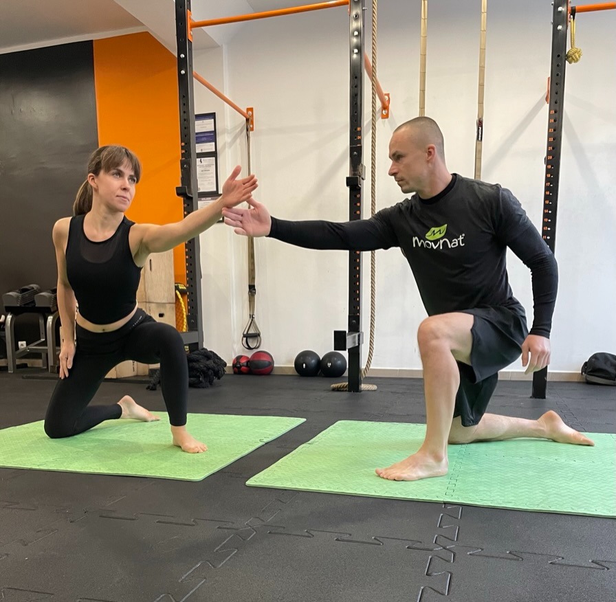 Kobieta i mężczyzna w strojach sportowych wykonują ćwiczenie rozciągające na zielonych matach w siłowni, z wyciągniętymi dłońmi ku sobie.