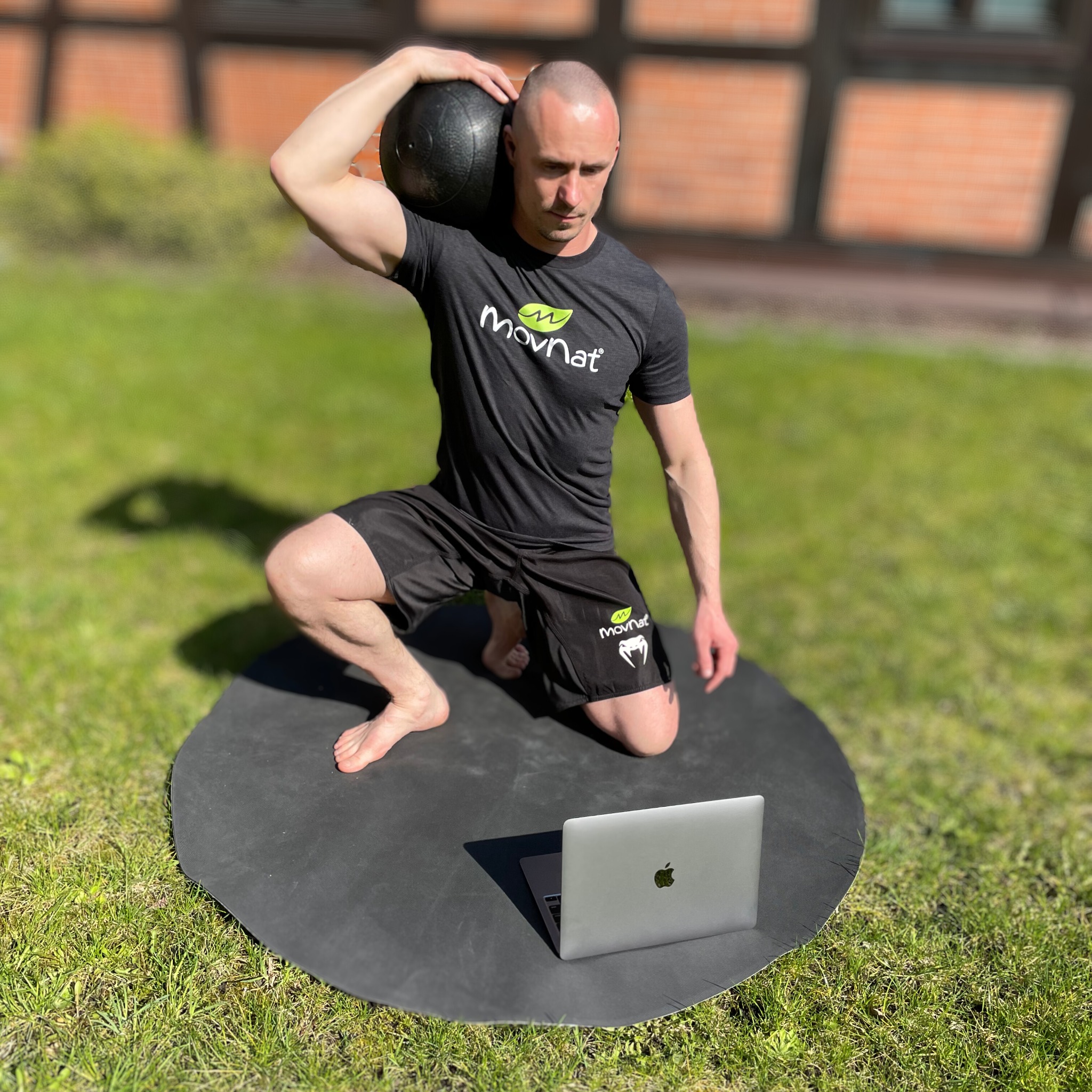 Mężczyzna w czarnym stroju sportowym klęczy na macie na trawie, trzymając czarną piłkę lekarską na ramieniu i patrząc na laptopa.