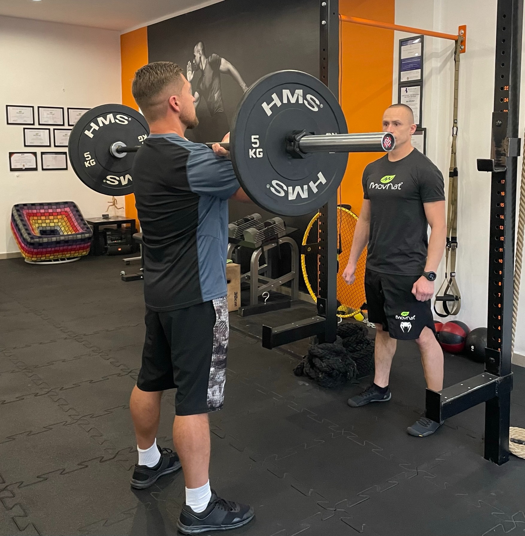 Mężczyzna ćwiczy podnoszenie sztangi pod nadzorem trenera w siłowni, widoczne obciążenia HMS 5kg, trener z ustnikiem zabezpieczającym i logiem Movnat na koszulce i spodenkach.