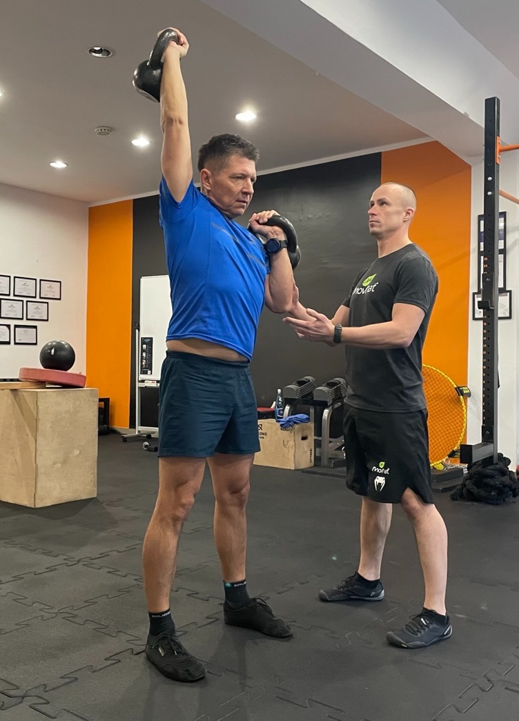 Mężczyzna w niebieskiej koszulce podnosi kettlebell nad głowę, instruktor asekuruje ramię; ćwiczenie siłowe w siłowni z pomarańczową ścianą.