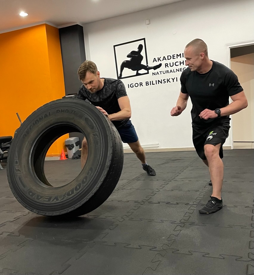 Dwóch mężczyzn ćwiczy z dużą oponą w siłowni, na ścianie logo 'Akademia Ruchu Naturalnego Igor Bilinskyi', jeden z mężczyzn w czarnej koszulce i spodenkach, drugi w czarnej koszulce i ciemnych...